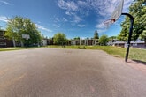 Located in Surrey, BC, this expansive communal courtyard serves as the central outdoor hub for a multi-building residential complex. The grounds are generously landscaped with well-maintained grass lawns and a variety of mature trees — including towering conifers and leafy deciduous trees — that provide ample shade and a sense of established, natural character throughout the space.

Paved pathways wind through the courtyard, connecting the various amenity areas and the surrounding three-storey residential buildings, which feature balconies overlooking the grounds. A full-sized basketball court offers recreational space, complemented by a dedicated children's playground area equipped with play structures set on wood chip surfacing. Seating is provided throughout, including wooden benches positioned along the pathways for residents to enjoy the outdoors.

A detached utility or storage building with garage access sits at one end of the courtyard, adjacent to raised garden beds where residents can cultivate plants and vegetables. The overall layout encourages outdoor activity and community interaction, with open green spaces providing room to gather or relax. Under sunny skies, the mature tree canopy, open lawns, and accessible amenities combine to create a welcoming and functional shared outdoor environment.