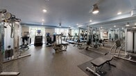 Located at 6205 Carnegie Blvd in Charlotte, NC, this well-appointed fitness center offers a comprehensive workout environment within a single, open-plan floor. The space is thoughtfully organized to accommodate a wide range of exercise needs, from cardiovascular training to strength and resistance work.

The cardio section features a row of treadmills, elliptical machines, and stair climbers, each equipped with individual display screens. Large windows along the perimeter flood the space with natural light and provide views of the surrounding greenery, creating an inviting atmosphere for exercise.

The strength training area is equally well-equipped, with multiple cable and pulley machines, a cable crossover station, and resistance training rigs. A dedicated free weights section includes a full rack of dumbbells, medicine balls in varying weights, a flat bench, and a barbell station, catering to all levels of fitness.

Underfoot, durable rubber flooring spans the entire space, providing safety and comfort during workouts. Floor-to-ceiling mirrors line portions of the walls, enhancing the sense of openness and allowing users to monitor their form. Recessed lighting and industrial-style pendant fixtures provide consistent illumination throughout. A small amenity station with shelving and a water dispenser adds a practical convenience. The facility is accessed through double glass doors and maintains a clean, modern aesthetic throughout.