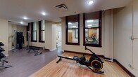 Located at 111 N Third Ave W in downtown Duluth, Minnesota, this fitness facility occupies a single-floor space within a historic building that seamlessly blends original architectural character with modern amenities. Exposed brick columns and tall, dark-framed windows preserve the building's heritage, while warm yellow walls, updated drop ceilings with recessed panel lighting, and clean white trim create a bright and welcoming environment throughout.

The facility is organized across several interconnected areas. A dedicated cardio room houses a treadmill, elliptical trainer, and stationary cycle, all set on durable carpet flooring and illuminated by flush panel lighting. Adjacent to this is a strength training zone equipped with a multi-station cable machine, a tiered dumbbell rack with a full range of free weights, and a flat bench for versatile workouts.

A spacious open room provides a flexible area ideal for stretching, floor exercises, or group activities, featuring large wall-mounted mirrors that enhance the sense of space and allow for form checking. Warm hardwood flooring in the main area adds a refined touch, contrasting nicely with the carpeted exercise zones. A rowing machine is positioned here, offering a full-body cardio option.

Interior windows connect the various workout zones, promoting visibility and an open feel throughout. Classic wooden doors with transom windows add a distinctive historic charm to the entryways, completing the character of this well-appointed fitness space.