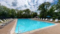 Located at Birch House in Douglassville, Pennsylvania, this outdoor recreational amenity area offers a well-appointed community pool and surrounding grounds designed for relaxation and leisure. The centerpiece is a generously sized outdoor swimming pool with crystal-clear blue water, framed by a wide concrete pool deck. The deck is lined with rows of white lounge chairs, providing ample space for sunbathing and poolside relaxation. Blue patio umbrellas are positioned throughout the deck and adjacent grassy areas, offering shaded seating options for guests seeking relief from the sun. A covered wooden gazebo sits at one end of the pool area, providing additional shaded space with seating and tables — ideal for casual gatherings. The facility is enclosed by a black iron perimeter fence, ensuring a secure and defined recreational space. Adjacent to the pool, a dedicated bocce ball court is set into a circular sand pad on a well-maintained grassy lawn, adding a classic outdoor game option for community members. The entire area is surrounded by mature trees that create a lush, green backdrop, enhancing the sense of privacy and natural ambiance. The setting offers an inviting, resort-like atmosphere suited for community recreation throughout the warmer months.