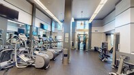 Located at 250 Congress Park Dr in Delray Beach, Florida, this well-appointed fitness center spans a single floor and is organized across two open workout areas. The space features impressively high ceilings that lend an airy, open atmosphere throughout, complemented by large floor-to-ceiling windows that flood the interior with natural light and offer views of the surrounding greenery. The flooring throughout is durable rubber gym matting, ideal for high-traffic exercise environments. The primary cardio area is lined with Precor treadmills and elliptical trainers, each equipped with individual display screens, positioned alongside full-wall mirrors and wall-mounted televisions to enhance the workout experience. Overhead fluorescent lighting ensures the space remains bright and functional at all times. The secondary area houses a multi-station cable strength machine, selectorized weight machines for targeted muscle training, and additional cardio equipment including upright bikes and a rowing machine. A built-in bench with cubbies provides a convenient space for personal belongings. Decorative pendant lighting and a consistent gray-and-white color palette give the facility a clean, modern aesthetic. The thoughtful layout and diverse equipment selection make this fitness center a comprehensive amenity for users of all fitness levels.