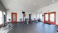 Located at 8181 Herald Cv in Blacklick, Ohio, this single-story fitness center offers a well-equipped workout space designed to support a range of exercise routines. The interior features clean white walls and a vaulted ceiling that lends a sense of openness and airiness to the room. Recessed lighting throughout ensures the space is bright and well-illuminated, complemented by natural light streaming in through windows along the exterior wall.

The durable dark rubber flooring covers the entire workout area, providing both safety and comfort underfoot. The space is outfitted with a multi-station cable weight machine, a flat weight bench, and a dumbbell rack stocked with a variety of free weights, catering to both strength training and toning workouts. A wall-mounted television is positioned for easy viewing during exercise sessions.

Access is provided through two red-framed doors — one with glass panel detailing — which add a bold accent to the otherwise neutral interior. The layout is thoughtfully organized to allow for free movement between equipment. Whether used for a focused strength session or a casual workout, this fitness center provides a functional and inviting environment for residents and visitors alike.