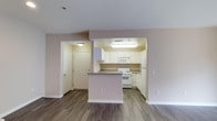 This single-floor residence offers a clean, open-concept layout that seamlessly connects the living and kitchen areas, creating a sense of spaciousness and ease of movement. The kitchen is well-appointed with white cabinetry, a full suite of white appliances including a refrigerator, electric range, and built-in microwave, complemented by granite-style countertops. A peninsula breakfast bar extends into the living space, providing additional prep space and casual dining options. The hardwood-style flooring runs throughout the main living area, offering both durability and a warm aesthetic. Neutral, light-toned walls contribute to a bright and airy atmosphere, enhancing the overall openness of the space. A textured ceiling adds subtle dimension overhead, while recessed and flush-mount lighting ensures consistent illumination throughout. Interior doors feature classic raised-panel detailing, adding a touch of traditional character to the home. The layout also includes what appears to be a dedicated closet or storage area near the entryway, offering practical organization solutions. Overall, this residence presents a functional and comfortable living environment with a cohesive, understated design aesthetic.