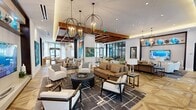 This expansive, single-floor amenity space offers a generously sized, open-plan layout designed for both socializing and relaxation. The interior is anchored by striking geometric-patterned hardwood flooring that flows seamlessly throughout, drawing the eye across each distinct zone of the space.

The lounge area is elegantly appointed with contemporary furnishings, including plush sofas, armchairs, and a large-screen television set into a built-in media wall adorned with decorative accents. Pendant chandeliers with orbital metal frames hang above, casting warm ambient light complemented by recessed ceiling lighting. Large floor-to-ceiling windows and glass-paneled entry doors flood the interior with natural light and provide clear sightlines to the outdoors.

A dedicated dining and event area features a long conference-style table with upholstered seating beneath an architecturally detailed coffered ceiling with exposed warm-toned wood beams. A built-in wet bar and cabinetry serve this zone, making it ideal for gatherings. Adjacent to this area, a shuffleboard table adds a recreational element alongside a billiards table, further enhancing the entertainment offerings.

Large-scale abstract artwork in bold, vibrant colors is displayed throughout, contributing to the curated, gallery-like atmosphere. Exposed white brick walls and a sophisticated neutral color palette with warm wood accents complete the refined, modern aesthetic of this well-appointed communal space.