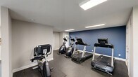 Located at Hamiltons Harbor in Lake Wylie, South Carolina, this community fitness center offers a generously sized, single-floor exercise space designed to support a comprehensive workout routine. The facility operates 24 hours a day, providing residents with flexible access at any time.

The interior features a clean, modern aesthetic with neutral gray walls, crisp white trim, and striking blue accent walls that define the free weights area. Durable rubberized flooring covers the entire space, providing both safety and comfort during exercise. Recessed and flush-mounted linear lighting fixtures illuminate the facility evenly throughout.

The cardio zone is outfitted with multiple treadmills, elliptical machines, and a stationary cycle, all positioned to take advantage of natural light from large windows and a sliding glass door that opens to an outdoor area. The strength training section includes a multi-station cable machine, a full rack of dumbbells, an adjustable weight bench, medicine balls, and a stability ball, offering a well-rounded selection for users of all fitness levels.

A large wall mirror in the strength area enhances the sense of space and allows for form monitoring. Additional doors lead to ancillary rooms, and the open layout ensures ample floor space for stretching or bodyweight exercises. Overall, the facility presents a bright, functional, and well-maintained environment for community fitness.