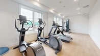 Located at 402 Halstead Ave in Harrison, NY, this fitness facility offers a clean, modern workout environment designed for both cardio and strength training. The gym features a light-filled interior enhanced by large windows along the exterior wall, allowing natural light to flood the space and provide views of the surrounding neighborhood — creating an energizing atmosphere for exercise.

The room is finished with light hardwood-style flooring that complements the crisp white walls and recessed ceiling lighting, giving the space a bright and open feel. The layout is thoughtfully organized, with cardio equipment prominently arranged near the windows, including a treadmill, elliptical trainers by Precor, and a Peloton stationary bike. A multi-functional cable machine and adjustable weight bench provide strength training options, while a dumbbell rack stocked with free weights and a stability ball round out the functional fitness offerings.

The ceiling features recessed lighting supplemented by HVAC vents, ensuring a comfortable environment year-round. A clearly marked exit door and dedicated wall panels reflect the facility's attention to safety and practicality. The overall ambiance is clean, uncluttered, and professional — making this gym a fully functional fitness space well-suited for a range of workout routines.