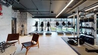 Located at 1000 Lake St in Oak Park, IL, this contemporary fitness center is designed to offer a comprehensive and visually engaging workout environment. The space is spread across a single floor and is divided into several distinct functional zones, each thoughtfully appointed with high-quality equipment and modern finishes.

The cardio area features a row of treadmills positioned directly in front of expansive floor-to-ceiling windows, flooding the space with natural light and offering unobstructed views of the outdoor courtyard. Light hardwood flooring runs throughout, complementing the industrial-style exposed ceiling with dark ductwork and sleek linear lighting.

The strength and functional training zone is equipped with Freemotion power racks, weight plates, kettlebells, and battle ropes, all set on rubber flooring for safety and durability. A hanging heavy bag adds a boxing element to the training repertoire.

The cycling and cardio studio area accommodates multiple stationary bikes and an elliptical, with large mirrored walls and pendant lighting creating an open, motivating atmosphere. Courtyard views from this section further enhance the workout experience.

A dedicated lounge area provides a space to relax and recover, featuring woven leather chairs, a marble-topped table, a wall-mounted television, and eclectic wall décor. The raw concrete walls and exposed overhead piping lend the space an industrial-chic aesthetic. Throughout, typographic graphic wallcoverings add a bold, urban character to the overall design.