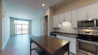 This well-designed single-floor unit offers a thoughtful layout that balances open living with practical functionality. The kitchen is a standout feature, outfitted with white shaker-style cabinetry, dark countertops, and a full suite of stainless steel appliances including a refrigerator, electric range, microwave, and dishwasher. Light wood-tone flooring runs throughout the main living areas, lending warmth and continuity to the space. A built-in pantry with wire shelving provides convenient additional storage just off the kitchen. The open living area is bright and airy, benefiting from large floor-to-ceiling windows and a sliding glass door that opens onto a private balcony with outdoor views. A dedicated laundry room houses a full-size side-by-side washer and dryer, complemented by overhead wire shelving for added utility. The bedroom is carpeted for comfort and features exposed ductwork along the ceiling, lending an industrial character to the space, along with a window that allows natural light. Adjacent to the bedroom is a generously sized walk-in closet with multi-level wire shelving and hanging rods. The full bathroom includes a single vanity with white cabinetry, a toilet, and a tub-shower combination, finished with wood-look tile flooring throughout.