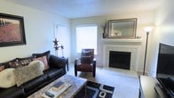 This single-floor space centers around a welcoming living room designed for comfort and functionality. The room features a classic white-manteled fireplace with light tile surround, serving as a natural focal point and adding warmth to the space. Above the mantel, a decorative framed mirror enhances the sense of depth and reflects natural light throughout the room.

The flooring is wall-to-wall carpet, providing a soft and comfortable feel underfoot, with tile extending around the fireplace hearth area. Two windows fitted with white horizontal blinds allow ample natural light to brighten the neutral, light-colored walls, contributing to an open and airy ambiance.

The living area comfortably accommodates seating, including a dark leather sofa and a rich brown accent chair, complemented by a marble-style coffee table. Decorative touches such as metal star sculptures, a tall vase with dried botanicals, and a patterned area rug add character to the space. The textured popcorn ceiling completes the traditional interior aesthetic of this cozy, well-appointed living space.