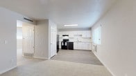 This single-floor residence features a practical, open layout that seamlessly connects the living and kitchen areas. The living space is finished with neutral-toned carpet, providing a comfortable and cohesive feel throughout. A ceiling fan with integrated lighting serves the main living room, adding both function and airflow. The kitchen is clearly defined by light wood-look vinyl flooring and is well-equipped with white shaker-style cabinetry, white subway tile backsplash, light-toned countertops, and a full suite of black appliances including a refrigerator, electric range with overhead ventilation, and dishwasher. A stainless steel sink is positioned beneath a window, allowing natural light into the cooking space. The bedroom areas continue the carpeted flooring and are illuminated by windows fitted with horizontal blinds for light control and privacy. The bathroom features a white vanity with ample cabinet storage, a large wall mirror, light countertops, and a shower-tub combination. Throughout the unit, neutral gray-white walls and white trim create a clean, consistent aesthetic. Flooring transitions clearly delineate the kitchen and bathroom from the carpeted living and sleeping areas.