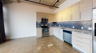 This single-floor urban loft combines industrial character with modern finishes throughout. The expansive main living area showcases polished concrete floors, soaring ceilings with exposed ductwork, and a bold track lighting system mounted on industrial-style rails. Large windows allow natural light to fill the open space, complemented by a distinctive curved wall accent and a striking red entry door that adds a pop of color to the aesthetic.

The kitchen is well-appointed and functional, featuring light wood cabinetry with sleek bar-pull hardware, a vibrant blue mosaic tile backsplash, and stone countertops. Stainless steel appliances — including a range, over-the-range microwave, dishwasher, and refrigerator — complete the space. The polished concrete flooring flows seamlessly from the main living area into the kitchen, reinforcing the loft's cohesive industrial design language.

The bathroom is thoughtfully designed with a floating wood-grain vanity, a vessel sink with a dark countertop, and large-format grey floor tiles. A combined soaking tub and shower is enclosed by a frameless sliding glass door and accented with dramatic dark mosaic wall tile, creating a refined, spa-like atmosphere. Recessed lighting and wall sconces provide a clean, well-lit environment throughout the bathroom.