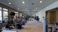 This fitness center offers a comprehensive workout environment designed to accommodate a wide range of exercise routines. The space is expansive, with a long, open floor plan that provides ample room for movement between equipment stations. Neutral-toned carpet tiles cover the flooring throughout, offering both comfort and durability underfoot.

A full wall of mirrors runs along one side of the facility, visually expanding the space and allowing users to monitor their form during workouts. Recessed lighting and ceiling fans are evenly distributed overhead, ensuring consistent illumination and airflow throughout. Large windows along one wall invite natural light and provide views to the exterior, contributing to an open and energizing atmosphere.

The cardio section is well-stocked with elliptical trainers, treadmills, and rowing machines, all from reputable brands such as Precor. A rack of free weights and medicine balls offers options for functional and strength training. The dedicated strength training area features cable machines and weight benches, providing a full suite of resistance equipment. A wall-mounted flat-screen television adds convenience for users during extended workout sessions. The facility is clean, well-maintained, and thoughtfully arranged to maximize both usability and safety.