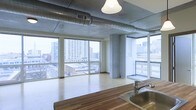 This single-floor space showcases a compelling blend of industrial and contemporary design elements. The open-plan layout is defined by its floor-to-ceiling windows on multiple walls, flooding the interior with natural light and offering expansive panoramic views of the surrounding urban skyline and streetscape below, suggesting an elevated position within a larger building.

The ceiling retains its raw, industrial character with exposed concrete and prominent silvery ventilation ductwork running horizontally across the space, lending an authentic loft aesthetic. A single pendant light fixture hangs centrally, complementing the industrial theme.

Light-toned hardwood flooring runs throughout the main living area, providing a warm contrast to the cooler concrete and metal elements overhead. Structural concrete columns punctuate the open floor plan, adding to the architectural character of the space.

The kitchen area is positioned at the foreground and features a warm wood-toned countertop and a stainless steel sink, offering a functional workspace that integrates naturally into the open layout. The overall ambiance is airy, modern, and distinctly urban, making strong use of natural light and city views as key design features.
