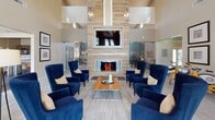 This single-floor community clubhouse offers a thoughtfully designed gathering space that balances elegance with functionality. The centerpiece of the facility is a grand lounge area featuring soaring ceilings, a striking floor-to-ceiling tiled fireplace, and a wall-mounted television framed in decorative tile. Six plush navy blue wingback chairs surround a sleek coffee table, creating an inviting focal point bathed in natural light from large windows. Elegant pendant chandeliers complete the sophisticated atmosphere.

Adjacent to the main lounge is a comfortable media room with expansive windows overlooking an outdoor patio area, furnished with sectional seating, accent chairs, and a large flat-screen TV. A cozy dining nook with a wood table, leather chairs, and game-themed wall art provides a casual retreat.

The modern kitchen features dark upper cabinetry, light lower cabinets with dark countertops, a tiled backsplash, stainless steel appliances including a French door refrigerator, and pendant lighting over a breakfast bar with metallic stools.

A glass-walled business center with multiple computer workstations and ample natural light from floor-to-ceiling windows serves as a productive workspace. Glass-enclosed private meeting rooms and a welcoming entry corridor with dark hardwood flooring and lush greenery round out this well-equipped community space.