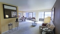 This well-appointed single-floor residence offers an expansive, open-plan layout that seamlessly integrates the living and dining areas into one cohesive, light-filled environment. Large floor-to-ceiling windows span multiple walls, flooding the interior with abundant natural light and framing impressive panoramic views of the surrounding urban skyline — a defining architectural feature of the space.

The living area is thoughtfully arranged with a circular glass-top coffee table as a central focal point, complemented by plush seating and warm accent pillows in earthy tones. Soft, neutral carpeting extends throughout, adding comfort underfoot and contributing to the space's overall warmth. The dining area is positioned near the windows, taking full advantage of the city views, and is furnished with a round table and cushioned chairs that invite casual yet refined gatherings.

Warm cream and yellow-toned walls enhance the sense of brightness and openness, while decorative accents — including a tall foliage arrangement, a large wall mirror, and curated tabletop pieces — add depth and personality to the space. The high, smooth ceilings further amplify the sense of volume and airiness throughout. Overall, the space strikes a balance between elegant design and comfortable urban living.