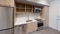 This single-floor space offers a clean, contemporary design characterized by neutral tones, grey hardwood-style flooring, and a practical open layout. The living area is bright and inviting, benefiting from large windows with dark frames that allow natural light to fill the room, offering views of the surrounding urban environment. The walls are finished in a soft white palette, complementing the modern aesthetic throughout.

The bedroom area is generously proportioned, featuring a closet and ceiling-mounted HVAC vents, ensuring year-round comfort. A structural soffit detail adds architectural interest to the space.

The kitchen is efficiently designed with light wood cabinetry, open upper shelving, white tile backsplash, and white quartz-style countertops. Stainless steel appliances include a dishwasher, electric range, and over-the-range microwave, providing a fully functional cooking environment.

The bathroom is finished with neutral tile flooring, a wood-toned vanity with an undermount sink, white countertop, a large mirror, and a glass-enclosed shower, all complemented by chrome fixtures. The overall ambiance of the space is clean, modern, and move-in ready.