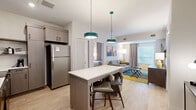 Located in Tucson, AZ, this 2-bedroom, 1-bathroom residence offers a thoughtfully designed single-floor layout that maximizes comfort and functionality. The open-concept kitchen and living area serves as the heart of the home, featuring light wood-style flooring, crisp white walls, and a central kitchen island with seating — illuminated by distinctive teal pendant lights. The kitchen is fully equipped with stainless steel appliances, including a full-size refrigerator, range, and microwave, complemented by taupe cabinetry with glass-front upper panels and a clean white countertop. The living room flows naturally from the kitchen, offering a wall-mounted flat-screen TV, a media console, and colorful Southwestern-inspired artwork that echoes the Tucson locale. Natural light enters through large windows dressed with blinds and neutral drapes. Both bedrooms are comfortably sized, each featuring warm wood-toned headboards, ample closet and shelving storage, and large windows. The primary bedroom includes a wall-mounted TV and full dresser. The bathroom is finished with wood-look tile flooring, a combined tub-shower, a wood vanity with open shelving, white countertops, and an illuminated mirror.