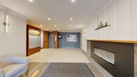 This residential building lobby presents a well-designed entrance space that balances contemporary finishes with warm architectural details. Large-format polished tiles span the floor throughout, reflecting the recessed pot lighting above and contributing to a bright, clean aesthetic. A central entry corridor leads from the glazed front doors, which frame views of the exterior brick archway and landscaped surroundings, allowing natural light to filter into the space.

To one side, a built-in linear fireplace is set within a white-panelled surround topped with a natural wood mantle, accented by a decorative sculptural piece mounted above. Adjacent to the fireplace, a comfortable seating area with neutral upholstered armchairs and a small side table provides a functional waiting space for residents and guests.

On the opposite side, a prominent wall of warm horizontal wood slats houses a large bank of individual resident mailboxes, neatly organized and well-lit. This feature wall adds texture and a natural warmth that contrasts effectively with the surrounding white and grey tones. Elevator access is centrally located at the rear, flanked by the same warm wood cladding, with a directory panel and small monitor nearby. The overall ambiance is modern, polished, and inviting.