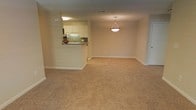 This single-floor residence offers a welcoming and functional layout designed with comfort and practicality in mind. The main living area is spacious and open, finished with plush wall-to-wall carpeting in a warm beige tone that adds softness underfoot and contributes to the room's cozy ambiance. Neutral-painted walls provide a clean, versatile backdrop that accommodates a variety of décor styles.

A standout feature is the pass-through kitchen window, which creates a natural connection between the kitchen and the living area, making the space feel open and social. The kitchen is equipped with white upper cabinetry, a sink with a green tile backsplash, and a breakfast bar-style counter, offering both storage and casual dining options. A decorative pendant light fixture hangs above the living area, providing a subtle focal point and warm overhead illumination.

Additional details include white baseboard trim throughout, a flush ceiling vent, and interior doors finished in a clean white — all contributing to a cohesive, tidy aesthetic. The overall atmosphere is calm and neutral, making it a practical and inviting space well-suited for everyday living.