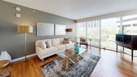 This single-floor residence offers a thoughtfully designed layout that balances comfort and functionality. The living area serves as the centerpiece of the space, featuring warm hardwood flooring, soft sage-green accent walls, and floor-to-ceiling windows that flood the room with natural light and provide views of the surrounding greenery. A wall-mounted television and a live-edge console table add character to a secondary living zone, which also includes convenient in-unit stacked laundry.

The kitchen is well-equipped for cooking enthusiasts, featuring warm wood cabinetry, dark granite countertops, and a full suite of stainless steel appliances including a gas range, built-in microwave, and refrigerator. Light tile flooring ties the space together cleanly.

The bedroom is a calm retreat with plush carpeting, soft neutral tones, and a large window offering leafy outdoor views. Ample storage and a spacious layout accommodate generous furnishings comfortably.

The bathroom is finished with light stone-effect tile throughout, a soaking tub with a shower and curtain, warm wood upper cabinetry, and a clean, minimalist vanity — creating a relaxing, functional space.