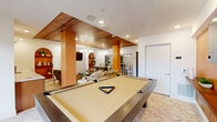 This well-appointed community lounge and game room offers a thoughtfully designed social space that balances recreational amenities with comfortable gathering areas. The room is anchored by a full-size pool table positioned beneath a distinctive wood-paneled ceiling inset, which adds warmth and visual depth to the open layout. Recessed lighting and a mid-century modern-style globe chandelier with brass accents provide layered illumination throughout the space.

The lounge area features a cushioned sofa and accent chairs arranged around a glass-top foosball table, creating an inviting hub for casual socializing. An arched built-in shelving unit with decorative accents adds architectural character to one wall, while a wall-mounted flat-screen TV is set against a bold orange accent wall above a wood credenza, making it a natural focal point for entertainment.

A compact wet bar with cabinetry and a small refrigerator is conveniently integrated into the space, ideal for hosting. Bold orange structural columns punctuate the room and complement the warm, earthy color palette. Large windows along one wall allow natural light to fill the space while offering views of the surrounding exterior. Patterned carpeting throughout ties the design together, and direct exterior door access adds to the room's functionality as a shared amenity space.