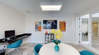 This single-floor space showcases a clean, modern aesthetic defined by crisp white walls and a neutral tiled floor laid in a subtle geometric pattern. The dining or gathering area is a notable focal point, centered around a sleek white round table paired with teal upholstered chairs that introduce a bold pop of color to the otherwise neutral palette. A wall-mounted flat-screen television serves as a functional centerpiece, flanked symmetrically by two large abstract paintings rendered in vivid oranges, blues, and whites, adding an artistic and dynamic energy to the space. A compact open shelving unit beneath the television provides additional display and storage. Overhead, a large flush-mounted panel light fixture delivers even, bright illumination throughout the area. A dark countertop surface is partially visible along one wall, suggesting additional utility space nearby. A vase of yellow orchids on the table adds a lively natural accent. The overall ambiance is contemporary and well-curated, combining bold color accents with minimalist architectural finishes to create a welcoming, visually engaging environment.