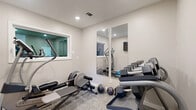 This dedicated fitness room is thoughtfully designed to maximize functionality within a compact footprint. The space features neutral light gray walls paired with low-pile carpet flooring in a subtle pattern, creating a clean, professional gym aesthetic. Recessed ceiling lighting provides consistent, even illumination throughout the room, complemented by a window that offers a view into an adjacent interior space, adding visual openness.

The room is equipped with a well-rounded selection of fitness equipment, including a Technogym elliptical machine and a rowing/ab bench for cardio and core training. A multi-tier dumbbell rack holds a complete set of hex dumbbells, while medicine balls and balance equipment are also present for functional training. Two large wall-mounted mirrors along one wall enhance the sense of space and allow users to monitor their form during workouts. A wall-mounted flat-screen television is positioned for easy viewing from multiple points in the room.

A standard entry door with a window provides natural light and access to the outdoors. A wall-mounted speaker is also visible, supporting audio during workouts. The overall design is efficient and purposeful, delivering a fully functional private fitness environment.
