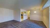 This single-floor space offers a practical and functional layout centered around an open-plan living and dining area. The living space is finished with wall-to-wall carpeting in a neutral grey tone, providing a comfortable and cohesive feel underfoot. A pendant light fixture hangs over the dining area, offering focused overhead illumination that complements the overall warmth of the space.

The kitchen is partially separated from the main living area by a dividing half-wall, which incorporates a pass-through counter — a feature that maintains a sense of openness while defining each functional zone. The kitchen itself is finished with light tile flooring and features warm-toned wood cabinetry, a white refrigerator, and under-cabinet lighting that brightens the work surfaces. A mirrored or window element above the sink adds depth to the kitchen space.

A hallway visible from the main living area suggests access to additional rooms beyond, contributing to the unit's overall sense of depth. The neutral wall tones throughout create a clean, versatile backdrop. Recessed and ceiling-mounted lighting provides consistent illumination across all areas of this well-organized, single-level layout.