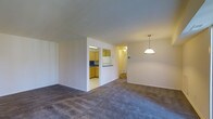 This single-floor space offers a practical and functional layout centered around an open-plan living and dining area. The living space is finished with wall-to-wall carpeting in a neutral grey tone, providing a comfortable and cohesive feel underfoot. A pendant light fixture hangs over the dining area, offering focused overhead illumination that complements the overall warmth of the space.

The kitchen is partially separated from the main living area by a dividing half-wall, which incorporates a pass-through counter — a feature that maintains a sense of openness while defining each functional zone. The kitchen itself is finished with light tile flooring and features warm-toned wood cabinetry, a white refrigerator, and under-cabinet lighting that brightens the work surfaces. A mirrored or window element above the sink adds depth to the kitchen space.

A hallway visible from the main living area suggests access to additional rooms beyond, contributing to the unit's overall sense of depth. The neutral wall tones throughout create a clean, versatile backdrop. Recessed and ceiling-mounted lighting provides consistent illumination across all areas of this well-organized, single-level layout.