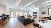 This thoughtfully designed single-floor space offers an open, versatile layout that seamlessly blends comfort and functionality. The expansive main area is bathed in natural light from a striking row of skylights overhead, which complement the warm-toned hardwood flooring and wood-framed windows found throughout. Crisp white walls provide a clean backdrop that enhances the bright, airy ambiance of the entire space.

The lounge area serves as a central gathering point, anchored by a large sectional sofa, mid-century modern accent chairs, and a sleek wall-mounted fireplace with an integrated television above — ideal for relaxed socializing or informal meetings. Adjacent to this is a flexible dining and collaborative zone furnished with a mix of high-top tables and standard-height tables, accommodating a variety of group sizes and uses. Large sliding glass doors and wide windows offer views of the surrounding greenery, connecting the interior to the outdoors.

A dedicated nook features a bookshelf, artwork, and additional seating, providing a quieter retreat within the open floor plan. The fully equipped kitchen rounds out the space with stainless steel appliances, a large island with bar seating, glossy white upper cabinetry, open wood shelving, and a dark stone countertop — designed for both everyday use and hosting.