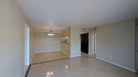 This single-floor residence offers an open, connected layout that combines the living and dining areas into one cohesive space. The flooring throughout is composed of large, light-toned ceramic tiles accented with a darker border detail, adding a subtle decorative element to the main living area. The dining space is defined by a modest chandelier overhead, providing a clear sense of separation from the living zone without the use of walls.

A pass-through counter connects the main living area to the kitchen, which features dark cabinetry and a black refrigerator, providing a functional and efficient workspace. The kitchen is compact yet well-organized, with its own overhead lighting.

The walls throughout are finished in a neutral, warm beige tone, contributing to a clean and understated ambiance. White baseboards frame the room and complement the light ceiling. Overhead HVAC venting is integrated into the ceiling, ensuring climate control throughout the space.

A doorway off the main area leads to additional rooms, suggesting a practical and straightforward floor plan designed for everyday living. The overall atmosphere is simple, bright, and easy to maintain, making it a functional and livable space.