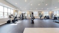 Located on S 60th St in Rogers, Arkansas, this single-floor fitness center offers a spacious and thoughtfully designed workout environment. The facility features soaring vaulted ceilings and recessed lighting throughout, creating a bright and open atmosphere. Large windows fitted with roller shades allow generous natural light to fill the space while providing privacy control, and ceiling fans help maintain comfortable airflow during workouts.

The main exercise floor is finished with durable rubber flooring and is well-organized across several distinct zones. The cardio area is outfitted with multiple treadmills and an elliptical machine, each positioned in front of wall-mounted flat-screen televisions for an engaging workout experience. A dedicated strength training zone includes a multi-station cable machine, rowing machines, and additional resistance equipment. Full-length mirrors line portions of the walls, enhancing the sense of space and aiding in form correction.

A separate functional training area features a free-standing punching bag, stability and medicine balls on a dedicated rack, and open floor space ideal for stretching or group exercise. A water fountain and informational signage are conveniently positioned near the entrance. The facility presents a clean, modern aesthetic with neutral warm-toned walls, making it a welcoming and fully functional space for a comprehensive fitness routine.