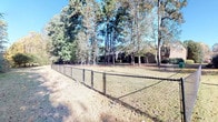This open-air lot offers a generous expanse of level ground framed by a mature, verdant landscape. The property is enclosed along key boundaries by a sturdy chain-link fence, providing clear definition of the space while maintaining an open, airy feel. Towering pine and hardwood trees line the perimeter, creating a natural canopy that offers both shade and a sense of privacy. The grassy interior ground cover stretches across the lot, transitioning at the edges into denser woodland with shrubs, vines, and native undergrowth — lending the space a secluded, natural character. One side of the property opens toward a well-maintained neighboring streetscape, where neatly trimmed boxwood hedges and manicured lawns are visible, suggesting a quiet, established residential setting. The wooded rear border provides a dense natural screen, while the open sky above ensures abundant natural light throughout the day. The lot's layout and generous proportions make it well-suited for a variety of uses, from new construction to recreational purposes. Overall, the property presents a peaceful, green environment with a strong sense of space, natural surroundings, and the potential to be shaped into a highly functional and appealing site.