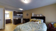 This single-floor studio is designed to maximize functionality within a compact footprint, seamlessly integrating sleeping, dining, and kitchen areas into one open layout. The sleeping area is anchored by a full-sized bed with a dark wood headboard featuring built-in shelving, providing convenient storage and display space. Warm tan walls contrast with light hardwood-style flooring, contributing to a comfortable and inviting ambiance throughout the space.

The kitchenette, positioned along one wall, is equipped with dark wood cabinetry, a compact refrigerator, a microwave, and a sink, offering essential cooking and food storage capabilities. A small dining area with a table and bar-style seating sits adjacent to the kitchenette, creating a practical spot for meals or studying. A textured ceiling with flush-mounted lighting provides consistent illumination throughout the room.

Overall, the studio is thoughtfully arranged to ensure each functional zone flows naturally into the next, making efficient use of available space while maintaining a cohesive, livable atmosphere suited for single-occupancy living.