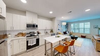 This well-designed single-floor residence offers a bright, contemporary living experience with a cohesive aesthetic throughout. The open-concept kitchen flows seamlessly into the living area, creating an inviting, connected space. The kitchen is outfitted with stainless steel appliances — including a refrigerator, range, and over-the-range microwave — complemented by crisp white cabinetry, a subway tile backsplash, and light countertops. A movable kitchen island with seating provides additional prep and dining space. Light-toned hardwood-style flooring runs throughout the main living areas, enhancing the sense of openness and continuity. The living room is anchored by a large window dressed with white blinds that allow generous natural light to fill the space. A lean wall-mounted shelving unit with an integrated desk area adds functional versatility to the corner of the room. The bedroom is a calm retreat, featuring a wide window for natural light, light-toned flooring, and access to a dedicated walk-in closet with built-in shelving and hanging rods. The bathroom presents a clean, all-white palette with subway-tiled tub and shower surround, a single vanity with under-cabinet storage, a wide mirror, and tile flooring — all contributing to a fresh, polished finish.