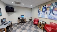 This single-floor business center and lounge is thoughtfully designed to serve as both a functional workspace and a relaxing communal area. The space features a distinctive checkered tile floor in muted earth tones that adds a stylish, dynamic character throughout. Recessed lighting set within a drop-panel ceiling provides bright, even illumination across the entire space.

The workstation area is equipped with multiple desktop computers, printers, and industrial-style wooden trestle desks paired with metal chairs, offering ample resources for productivity. A wall-mounted flat-screen television is positioned for easy viewing from the work area. Decorative artwork featuring bold "@" and "#" symbols adds a modern, tech-forward personality to this section.

The lounge area provides a welcoming retreat, furnished with a pair of red leather armchairs accented with nailhead trim, arranged around a game table. A beverage station along one wall offers a coffee maker and supplies for added convenience. Colorful, large-scale contemporary paintings — most notably a vivid street scene depicting pedestrians — enliven the neutral walls and contribute to an energetic yet comfortable ambiance. Together, the two areas create a well-rounded space that balances work and relaxation seamlessly.