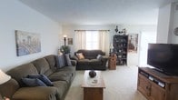 This two-floor home offers a warm and inviting atmosphere with a thoughtfully arranged interior. The living room serves as the heart of the main level, featuring wall-to-wall carpeting in a soft neutral tone that adds a sense of comfort underfoot. Two generously sized sofas in earthy brown tones are arranged to encourage conversation, complemented by a wooden coffee table at the center. Natural light filters through a window dressed with elegant cream curtains hung on a classic rod, brightening the room with a soft, airy glow. A tall floor lamp and a table lamp add layered lighting options for the evening hours. Along one wall, a dark wood bookcase displays a charming collection of figurines and decorative items, lending the room a personal and curated character. Framed artwork adorns the walls, contributing to the home's warm, lived-in aesthetic. A lush potted plant in the corner introduces a touch of nature to the space. The textured ceiling adds subtle visual interest overhead. The home's two-floor layout provides ample space for both daily living and private retreats, making it a well-rounded and functional residence suited for a variety of lifestyles.