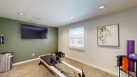 Located at 610 Paradise Ct in Greenwood, Indiana, this exercise room is a purpose-built fitness space designed to support focused, equipment-based workouts. The room features neutral-toned carpet tile flooring and a clean, modern aesthetic with a combination of soft green and light gray wall colors that create a calm, motivating atmosphere. Recessed lighting throughout the textured ceiling provides consistent, even illumination, while a standard window fitted with white horizontal blinds allows natural light to enter and offers a view of the surrounding area. A large wall-mounted flat-screen television serves as a functional centerpiece on the green accent wall, ideal for following guided workout sessions. The room's standout fixture is a full-length Pilates reformer positioned centrally, complemented by additional fitness accessories including a BOSU balance trainer and a multi-tier equipment rack stocked with exercise balls and foam rollers. Access to adjacent spaces is provided through two interior doors, with one featuring frosted glass panels, hinting at a broader facility beyond. Safety features including a fire extinguisher and exit signage are also present. The overall design balances practicality with a welcoming, studio-like ambiance suited for structured, low-impact exercise.