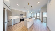 Located at 9590 E Northfield Blvd in Denver's Northfield neighborhood, this single-floor unit offers a thoughtfully designed layout with modern finishes throughout. The open-concept kitchen serves as the centerpiece of the living area, featuring soft gray shaker-style cabinetry with gold hardware, white quartz countertops, a full suite of stainless steel appliances, and a central island with an undermount sink and matte black faucet. Pendant lighting with warm brass accents illuminates the island, while recessed lighting and a ceiling fan enhance the adjacent living area, which opens to a glass door leading to an outdoor space. Light wood-style flooring runs throughout the main living and kitchen areas, contributing to a bright, airy atmosphere. The bedroom is carpeted for comfort and features a sliding barn door, clean white walls, and a large window that frames expansive city and skyline views. The bathroom is finished with large-format tile flooring and walls, a glass-enclosed walk-in shower, a gray vanity with gold hardware and a white countertop, a black-framed mirror, and gold light fixtures. Natural light filters in through a window, adding to the clean, contemporary feel. The unit combines functional design with upscale details throughout.