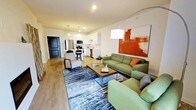 This single-floor space showcases an open-concept layout that seamlessly connects the living area and kitchen, creating a fluid and functional environment. The living area is anchored by a green upholstered sofa and matching accent chair, arranged around a sleek wood-and-metal coffee table atop a textured blue-grey area rug. A bold abstract painting in warm orange and red tones serves as a striking focal point on the main wall, complemented by a contemporary arc floor lamp that adds ambient lighting to the space.

The kitchen is well-appointed with white shaker-style cabinetry, stainless steel appliances including a refrigerator and microwave, and a white subway tile backsplash. A peninsula with bar seating provides a casual dining option and acts as a natural divider between the kitchen and living spaces. Pendant lighting above the peninsula adds a refined touch.

Throughout the unit, warm wood-tone laminate flooring provides continuity and contrast against the neutral white walls and dark-framed entry door. The overall ambiance is modern and inviting, with a carefully curated mix of textures and colors that create a cohesive, comfortable atmosphere.