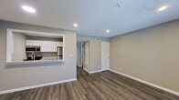 This single-floor unit offers a clean, modern design with a functional and open layout. The main living area is bright and welcoming, featuring warm hardwood-style flooring, neutral gray-toned walls, and recessed lighting throughout. Two windows along the front wall invite natural light into the space, creating an airy and comfortable atmosphere. Clean white baseboards provide a crisp contrast to the flooring and walls.

The kitchen is accessible via a pass-through counter that opens directly into the living area, enhancing the open-concept flow. It is outfitted with white shaker-style cabinetry, granite countertops, and stainless steel appliances including an electric range, over-the-range microwave, and refrigerator. The textured ceiling adds a classic touch present throughout the unit.

A hallway connects the living space to the additional rooms, including a full bathroom. The bathroom features a tub-shower combination with white tile surround, a toilet, a vanity with a light countertop, and a mirror. The same hardwood-style flooring extends into this space as well.

Overall, the unit presents a cohesive, move-in-ready aesthetic with consistent finishes, practical storage options, and a straightforward, well-organized floor plan suitable for everyday living.
