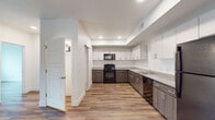 This single-floor residence showcases a clean, contemporary design with wood-look flooring and crisp white walls throughout, creating a bright and cohesive aesthetic. The spacious kitchen is a standout feature, offering an L-shaped layout with two-tone cabinetry — dark wood-grain lower cabinets paired with white upper cabinets — topped with granite-style countertops. Black appliances, including a gas range, microwave, dishwasher, and refrigerator, complete the kitchen alongside a double-basin stainless steel sink. Recessed lighting and a tray ceiling detail add dimension to the space. The open living areas are generously sized, with natural light flowing in through windows fitted with blinds. A dedicated laundry area with a washer and dryer is conveniently located within the unit. The residence includes two bathrooms: one featuring a granite-topped vanity with wood cabinetry and a tub-shower combination, and another with a matching vanity and access to a walk-in closet. The bedrooms are well-proportioned, each with wood-look flooring and windows that bring in natural light. Throughout the unit, the consistent use of neutral tones, modern finishes, and functional design elements create a polished and livable environment.