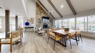 Located at 3304 Golden Rd in Tyler, TX, this community clubhouse offers an impressive and welcoming gathering space designed for both comfort and functionality. The interior is anchored by a dramatic vaulted ceiling with exposed dark wood beams and a ceiling fan, creating an airy, open atmosphere throughout the single-floor layout. A floor-to-ceiling stacked stone fireplace serves as the focal point of the living area, complemented by a wall-mounted television and decorative candle sconces, adding warmth and character to the space. Light wood-toned flooring runs throughout, enhancing the bright, neutral aesthetic. The open-plan layout accommodates both lounge seating arrangements and a dining area, making it well-suited for community gatherings and events. Large windows along the perimeter allow abundant natural light to pour in while offering views of the surrounding grounds. A mezzanine-level overlook adds architectural depth and visual interest to the space below, which features additional seating areas supported by structural columns. Outside, a generously sized freeform swimming pool is the centerpiece of the outdoor area, surrounded by a spacious concrete patio furnished with lounge chairs and umbrella-shaded tables. A covered porch adjacent to the building provides additional shaded outdoor seating, making the exterior an ideal extension of the clubhouse's social environment.