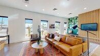 This single-floor space offers a thoughtfully designed interior that blends modern aesthetics with desert-inspired elements. The main lounge area features polished concrete flooring, a warm cognac leather sofa, and layered area rugs that add texture and color. A bold tropical-print accent wall anchors one corner of the room, complemented by vertical wood slat paneling and a built-in bench seating area with a large flat-screen display — ideal for gatherings or presentations. Multiple narrow windows line the walls, framing views of palm trees and cacti outside while filling the space with generous natural light. Live cactus plants and lush greenery are incorporated throughout, reinforcing the Southwestern character of the design. The kitchenette is outfitted with natural wood cabinetry, a crisp white countertop, a stainless steel sink, microwave, dishwasher, and a dark stacked-tile backsplash with open floating shelves styled with decorative ceramics and plants. The entry corridor features floor-to-ceiling glass partitions that open to a separate lounge nook with tufted booth seating, stone-textured accent walls, and additional outdoor-facing windows. Recessed lighting throughout keeps the overall ambiance bright, clean, and inviting.