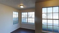 This single-story space showcases a bright, open interior defined by an abundance of natural light. The main living area features multiple large, multi-pane windows along two walls, allowing daylight to flood the room and offering views of the surrounding neighborhood. The dark-stained hardwood flooring provides a rich contrast against the warm beige walls, creating a balanced and inviting atmosphere. A flush-mount ceiling light fixture complements the textured popcorn ceiling overhead. White baseboards and window casings offer clean architectural detailing throughout. A partial wall or column element subtly divides the space, adding structural interest while maintaining an open feel. The neutral color scheme throughout makes the space versatile and well-suited to a variety of interior styles. Overall, the space conveys a comfortable, well-lit environment with classic residential finishes and a straightforward, functional layout.