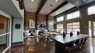 This single-floor communal space blends industrial and Texas-inspired design into a cohesive, inviting atmosphere. Soaring ceilings with dark exposed beams set an impressive tone throughout, while full-height exposed brick walls lend warmth and a rugged urban character to the interior. Polished concrete floors reflect light beautifully, enhancing the sense of openness across the generously sized room.

Natural light pours in through oversized industrial-style windows and garage-door-style glass panels that open the space toward an outdoor area, creating a seamless indoor-outdoor connection. Contemporary Sputnik-style chandeliers and pendant lighting provide atmospheric illumination throughout.

The lounge area features plush white wingback chairs, dark leather sofas arranged on patterned area rugs, and a round wooden coffee table — anchored by bold Texas-themed wall art, including a prominent "Texas Lone Star" poster. A wall-mounted flat-screen TV completes this relaxed seating zone.

A large communal dining table with modern chairs serves as a central gathering point, complemented by bar-height seating along one wall. A dedicated service counter area is equipped with commercial coffee and beverage appliances and dark granite countertops, decorated with decorative wine crest medallions overhead. Unique wall art, including a vintage Ford truck tailgate piece, adds eclectic character throughout the space.