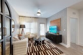 This thoughtfully designed single-floor residence blends modern aesthetics with bold decorative choices throughout its open and well-connected layout. The kitchen is a standout feature, showcasing grey cabinetry, granite countertops, stainless steel appliances including a French door refrigerator and range, a built-in wine rack, and a peninsula island with bar seating illuminated by pendant lighting. The living and dining areas flow seamlessly from the kitchen, featuring wood-look flooring, textured accent wallpaper, ceiling fans, and large windows with sheer curtains that invite ample natural light. A distinctive arched cabinet and bold patterned area rug add character to the living space, which also provides access to an outdoor area. The primary bedroom offers a serene retreat with clean white walls, wood-look flooring, and a spacious walk-in closet equipped with wire shelving and hanging rods. The en-suite bathroom features grey vanity cabinetry with granite countertops, a large mirror, and a glass-enclosed walk-in shower with marble-style tile. A second bedroom makes a bold statement with striking tiger-print wallpaper covering an entire accent wall, paired with a metal bed frame and warm ambient lighting. A separate dining nook completes the layout.