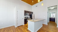 This single-floor unit showcases a thoughtfully designed open layout that combines modern finishes with striking architectural details. The living area immediately impresses with soaring ceilings accented by warm, natural wood paneling, ceiling fans, and pendant lighting that together create an inviting and airy atmosphere. Rich hardwood-style flooring runs throughout the entire space, providing a cohesive and polished look.

The kitchen is a standout feature, offering dark espresso cabinetry, granite-topped countertops, and a full suite of stainless steel appliances including a refrigerator, electric range, and microwave. A peninsula with a built-in sink and additional counter space extends into the living area, making it ideal for both cooking and entertaining.

Natural light pours in through a set of elegant double glass doors at the front entrance, which is fitted with transom windows above, further enhancing the bright and open feel of the living space.

The bathroom is well-appointed with a granite vanity, dark cabinetry matching the kitchen, hardwood-style flooring, and a full shower-tub combination. A dedicated laundry room provides added convenience with a full-size washer and dryer and overhead shelving. A private bedroom with a ceiling fan completes this well-rounded and functional space.