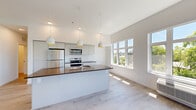 This thoughtfully designed single-floor home offers three bedrooms and two bathrooms within a bright, modern layout. The open-concept kitchen and living area serves as the heart of the home, featuring soaring vaulted ceilings, light hardwood-style flooring, and large windows that flood the space with natural light and frame views of surrounding greenery. The kitchen is well-appointed with stainless steel appliances — including a refrigerator, range, and microwave — sleek grey cabinetry, a white tile backsplash, and a substantial island with a dark countertop and undermount sink, illuminated by stylish pendant lights with warm gold interiors. The spacious primary bedroom receives generous natural light through oversized windows and includes dedicated closet space with built-in wire shelving. Additional bedrooms are equally bright, each with large windows overlooking tree-lined views. The bathrooms feature clean, modern finishes with dark countertops, undermount sinks, large mirrors, and tiled shower areas. A dedicated laundry room houses a full-size front-loading Whirlpool washer and dryer. Hallways are clean and well-lit, connecting all areas efficiently. Throughout the home, a consistent palette of white walls and light wood flooring creates a fresh, cohesive aesthetic.