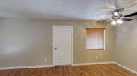 This single-floor space presents a straightforward, functional layout with a neutral and understated aesthetic. The main living area is finished with warm wood-look laminate or vinyl plank flooring that runs throughout, offering durability and a cohesive look underfoot. The walls are painted in a soft, warm greige tone that complements the flooring and contributes to a calm, neutral atmosphere throughout the space.

A classic white panel entry door serves as a focal point along the main wall, flanked by a window dressed with natural wood-toned horizontal blinds that allow for adjustable natural light. The window treatment adds a touch of warmth to the otherwise understated palette. Crisp white baseboards provide a clean contrast against both the walls and flooring, lending a finished, tidy appearance to the interior.

The ceiling features a textured popcorn finish, a common architectural detail that adds dimension overhead. Electrical outlets are conveniently positioned along the walls. The overall ambiance of the space is clean, bright, and move-in ready, offering a blank canvas that is well-suited for a variety of furnishing styles and personal touches.